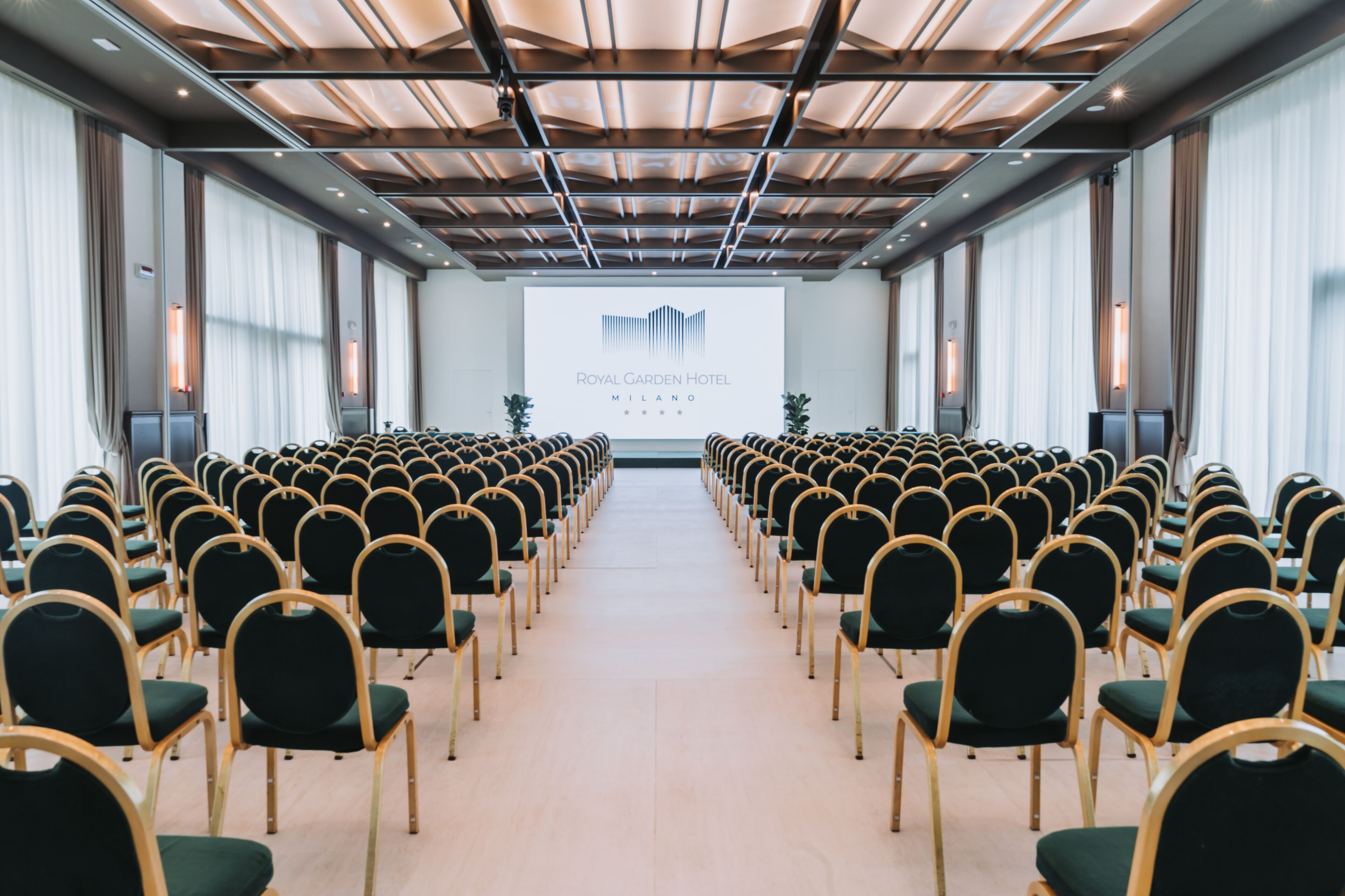 A conference room with rows of chairs facing a projection screen displaying the Royal Garden Hotel Milano logo.