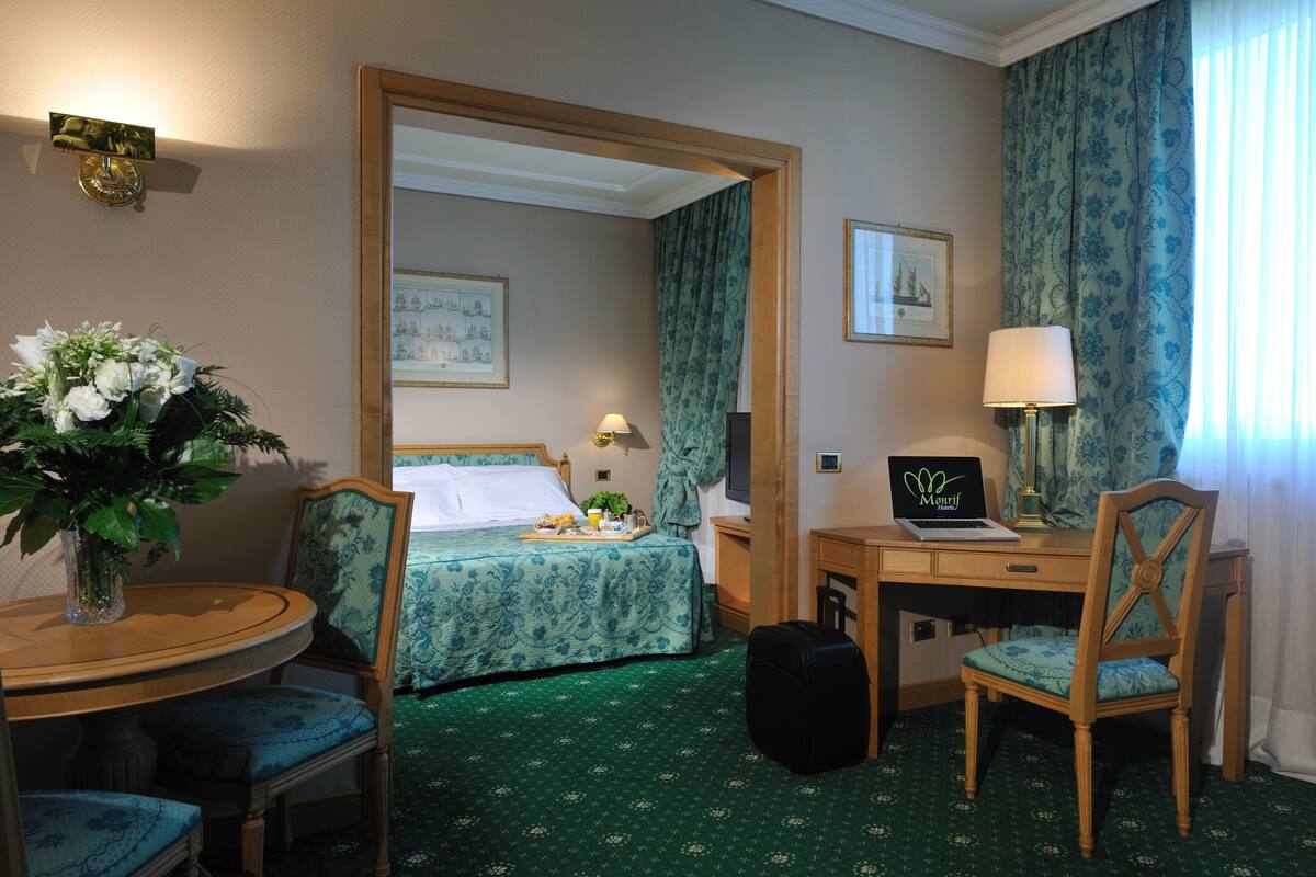 A hotel room with a bed, a desk with a laptop, a chair, a table with a vase of flowers, and a suitcase.