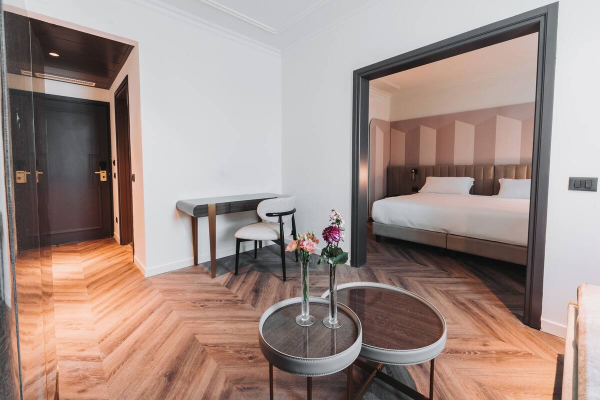 A hotel room with a bed, a small table with two vases, a chair, and a wooden floor.
