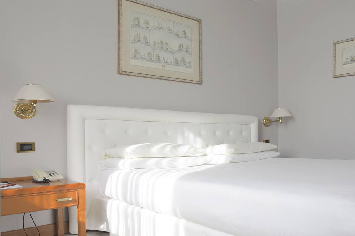 A neatly made bed with white linens, a wooden nightstand with a telephone, and two wall-mounted lamps.