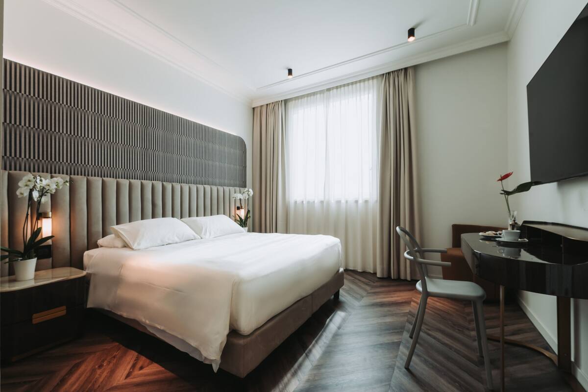 A modern hotel room with a large bed, a desk with a chair, a flat-screen TV, and a potted orchid.