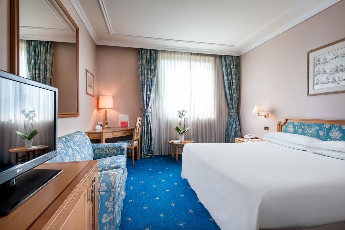 A hotel room with a bed, a desk with a lamp, a television, a chair, and a small table with a plant.