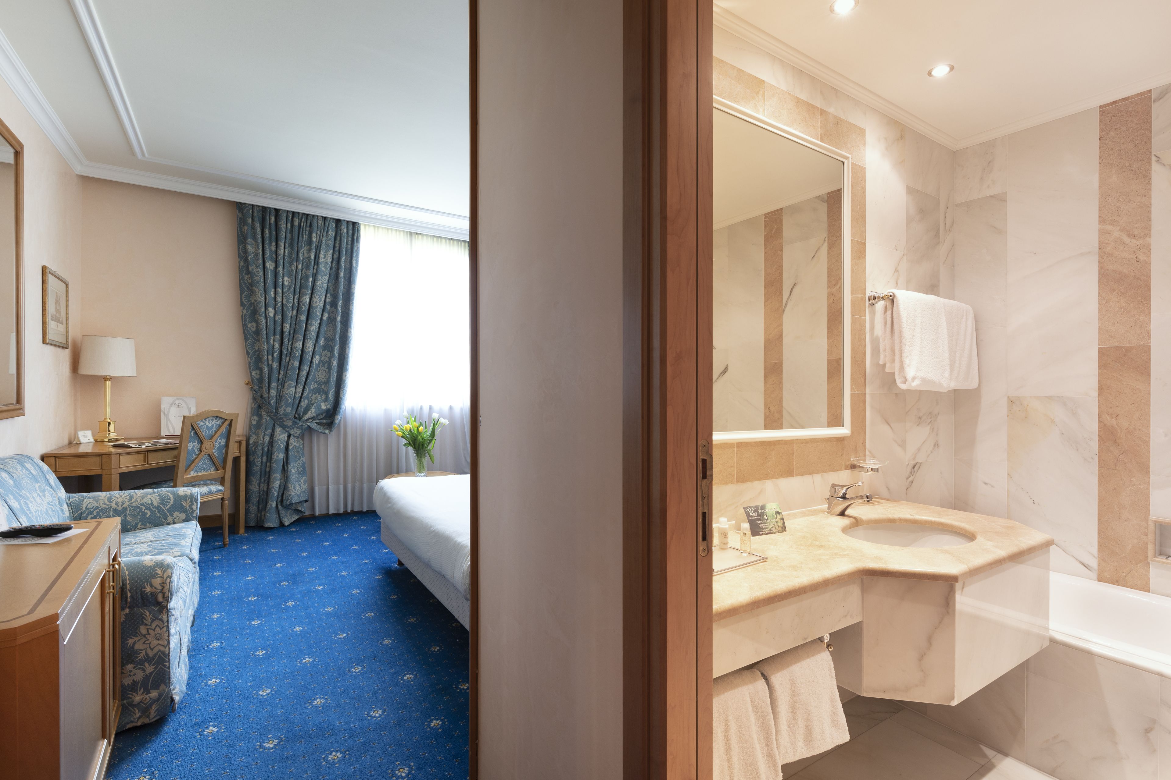 A hotel room with a bed, a desk, a chair, a desk lamp, a vase with flowers, and a bathroom with a sink, mirror, and bathtub.