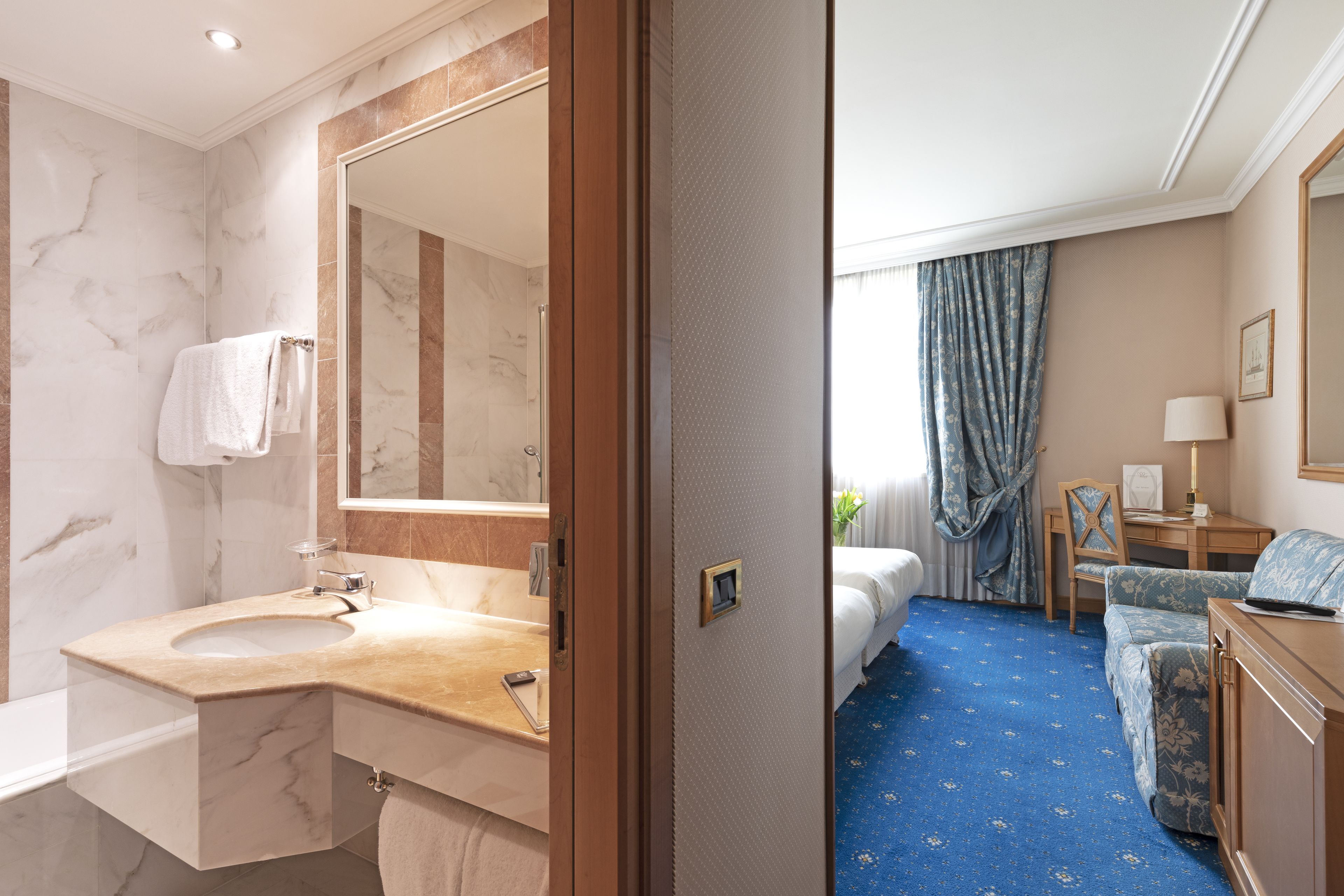 A hotel room with a marble bathroom, a bed with a blue floral carpet, and a desk with a lamp.