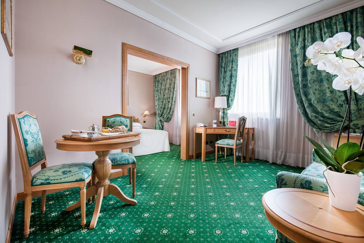 A hotel room with a bed, a desk with a chair, a round table with chairs, a potted plant, and a green patterned carpet.