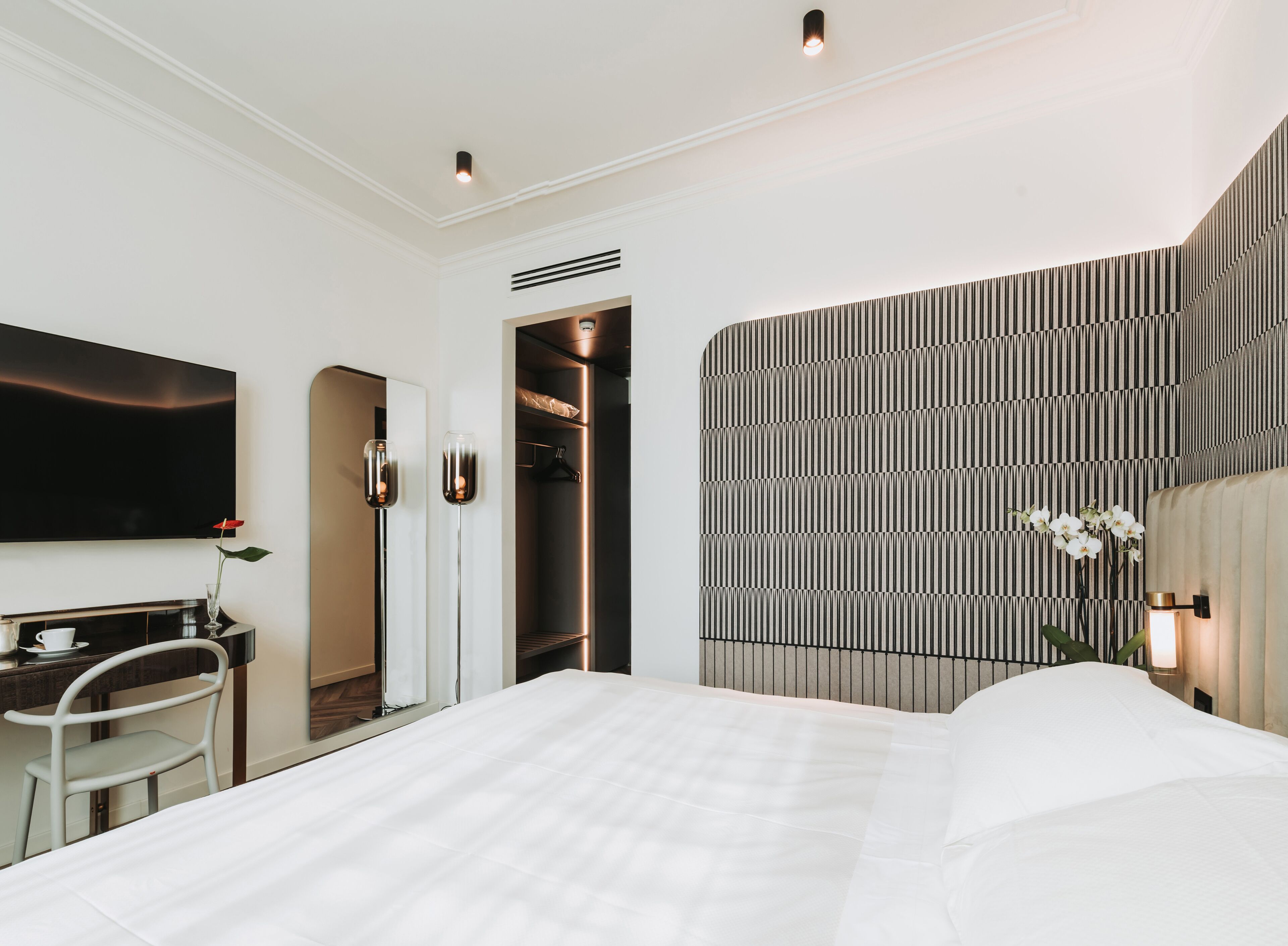 A modern hotel room with a large bed, a desk with a chair, a TV, and a wardrobe.
