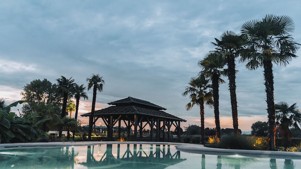 Una piscina cristalina con un pabellón en el centro, rodeada de palmeras y un cielo al atardecer.