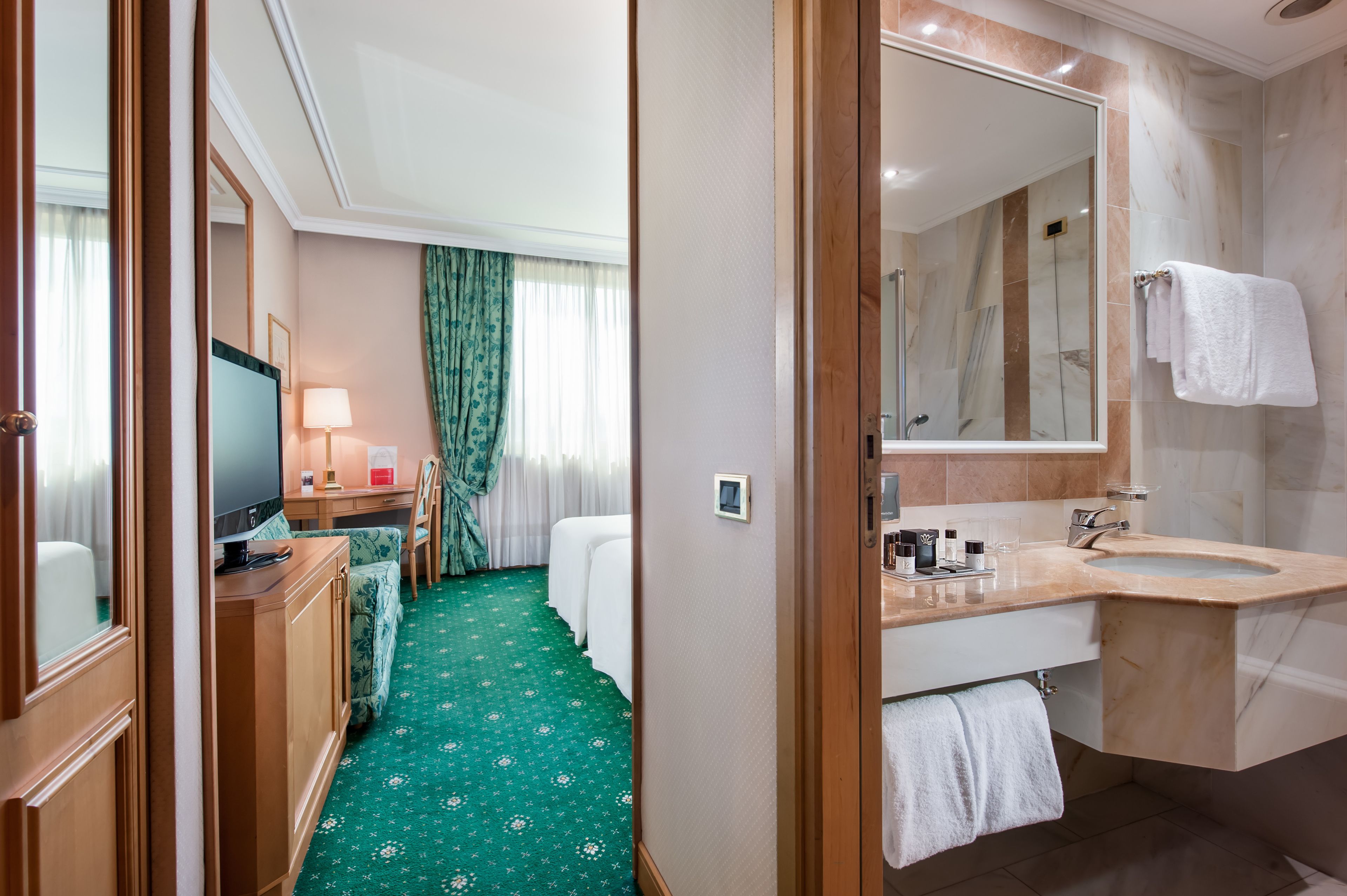A hotel room with a bed, a TV, a desk with a lamp, a bathroom with a sink and mirror, and a green carpet.