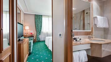 A hotel room with a bed, a TV, a desk with a lamp, a bathroom with a sink and mirror, and a green carpet.