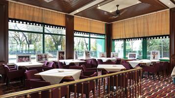 A restaurant with multiple tables and chairs, large windows with views of trees, and a patterned carpet.
