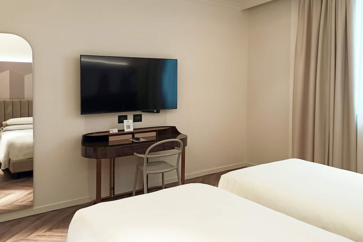 A hotel room with a bed, a desk with a chair, a flat-screen TV mounted on the wall, and a large mirror.