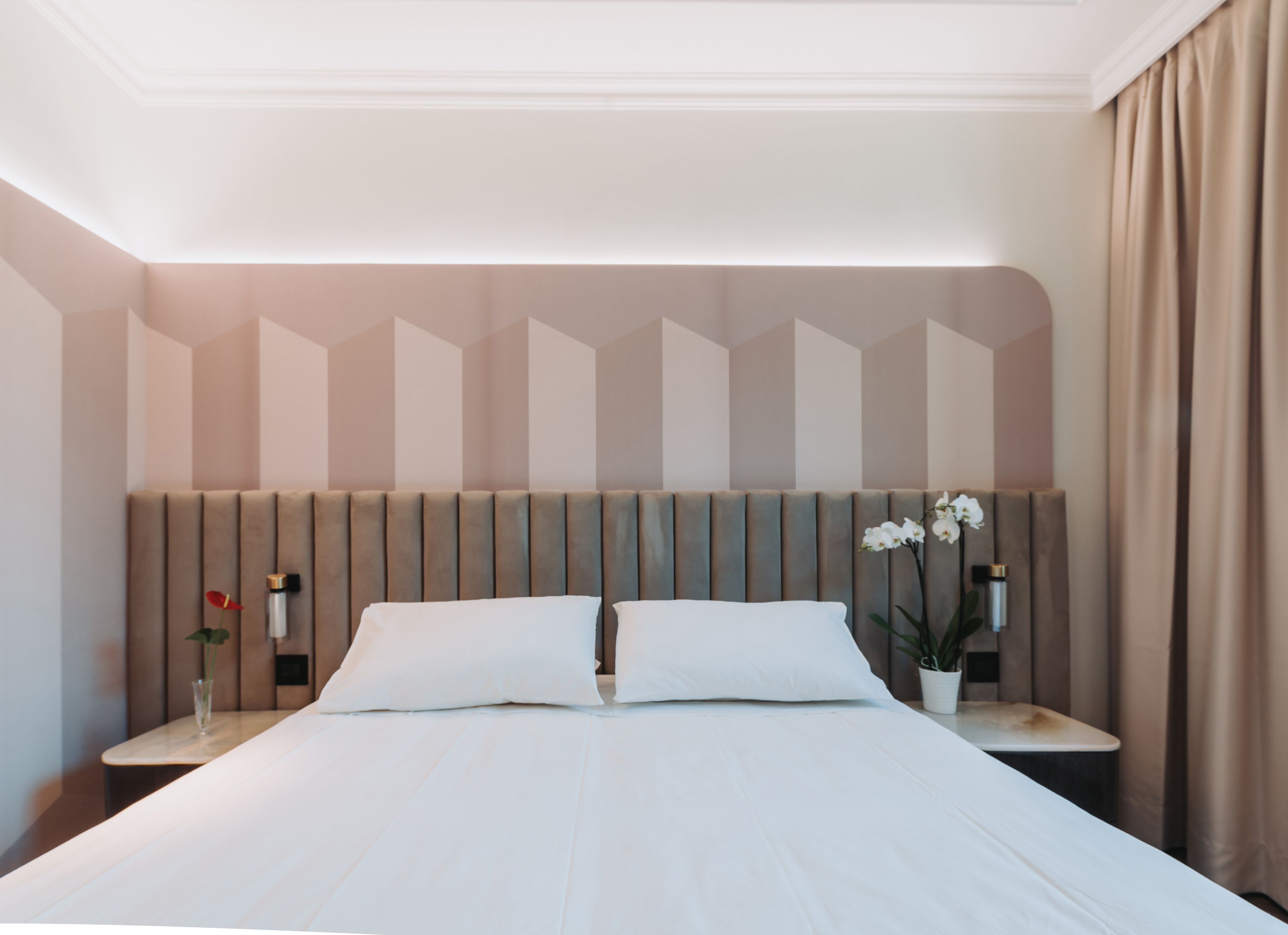 A modern hotel room with a large bed, a headboard with a geometric pattern, bedside tables with lamps, and a potted plant.