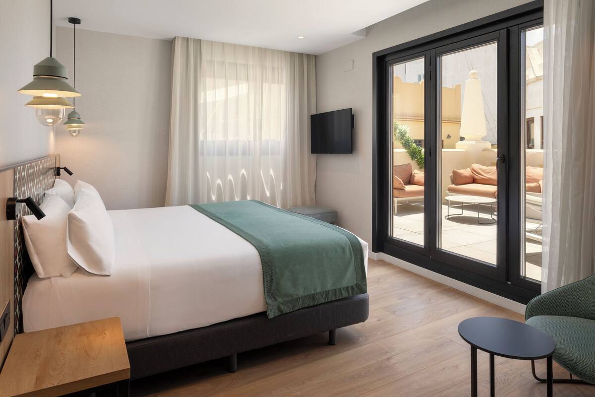 A hotel room with a bed, a TV, a balcony with seating, and a wooden floor.