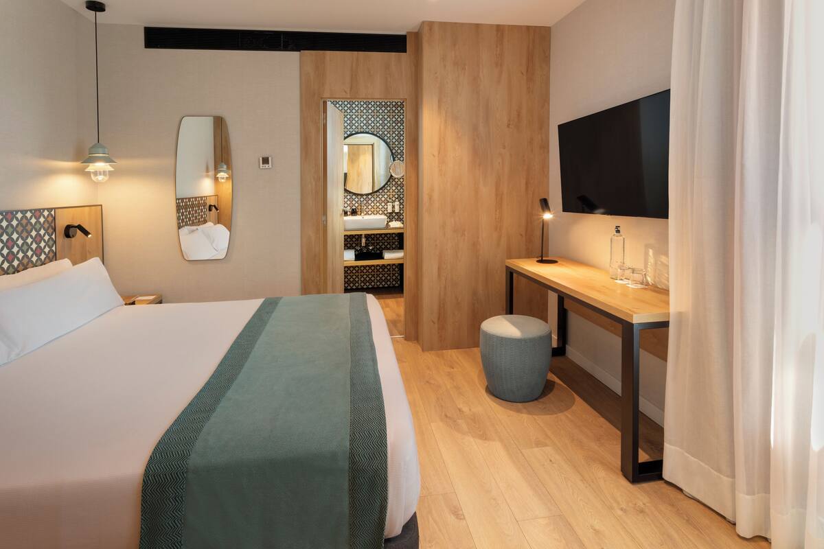 A modern hotel room with a large bed, a desk, a TV, and a bathroom with a mirror and sink.