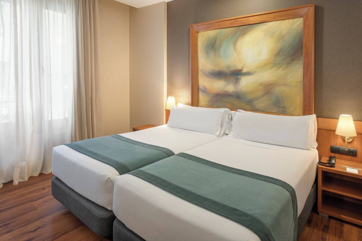 A double bed with two pillows, a green throw blanket, and a wooden nightstand with a lamp.