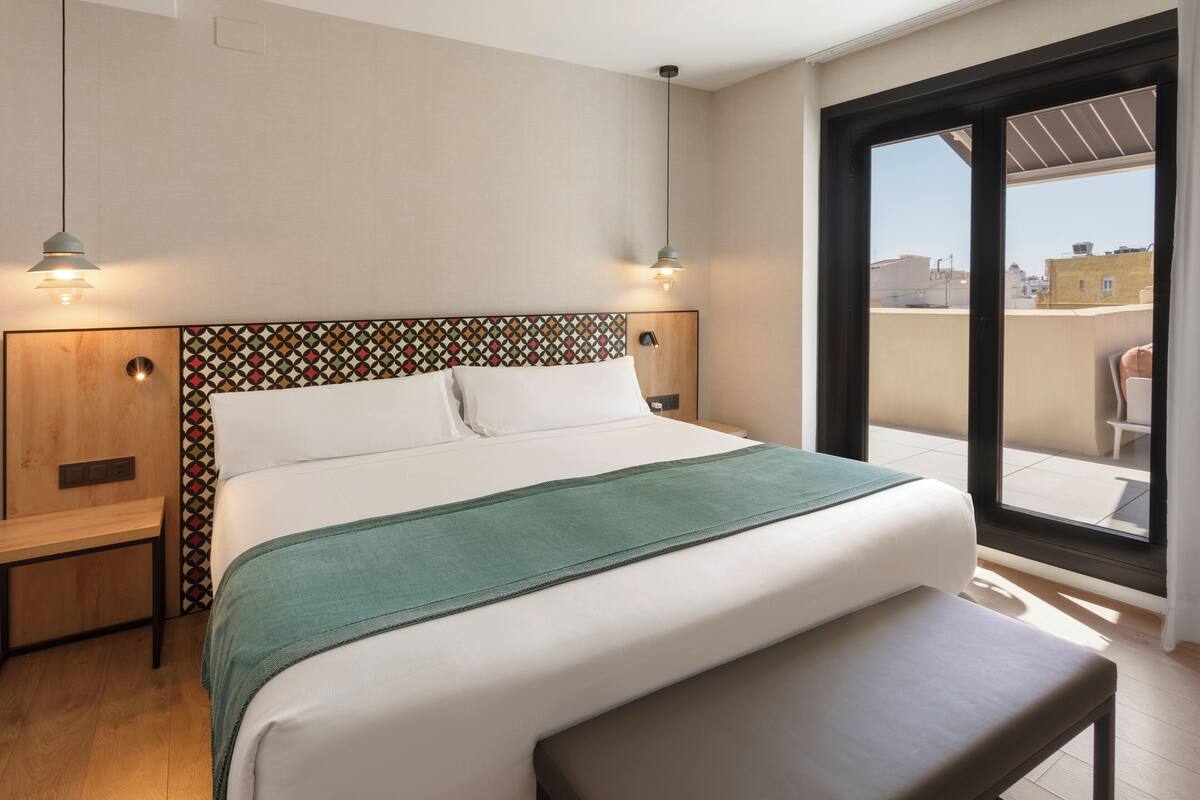 A neatly made bed with a green throw, wooden headboard with a geometric pattern, two bedside lamps, and a large window with a view of buildings.