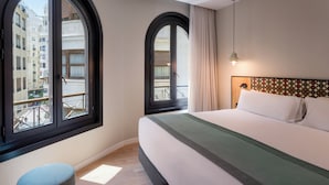 Habitación de hotel con una cama grande, un reposapiés redondo, una ventana con vistas a edificios y un cabecero estampado.