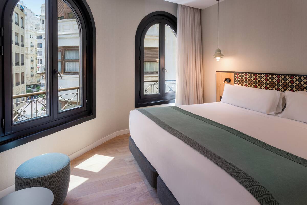 A hotel room with a large bed, a round ottoman, a window with a view of buildings, and a patterned headboard.