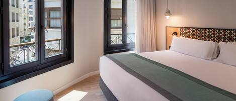 Habitación de hotel con una cama grande, un reposapiés redondo, una ventana con vistas a edificios y un cabecero estampado.