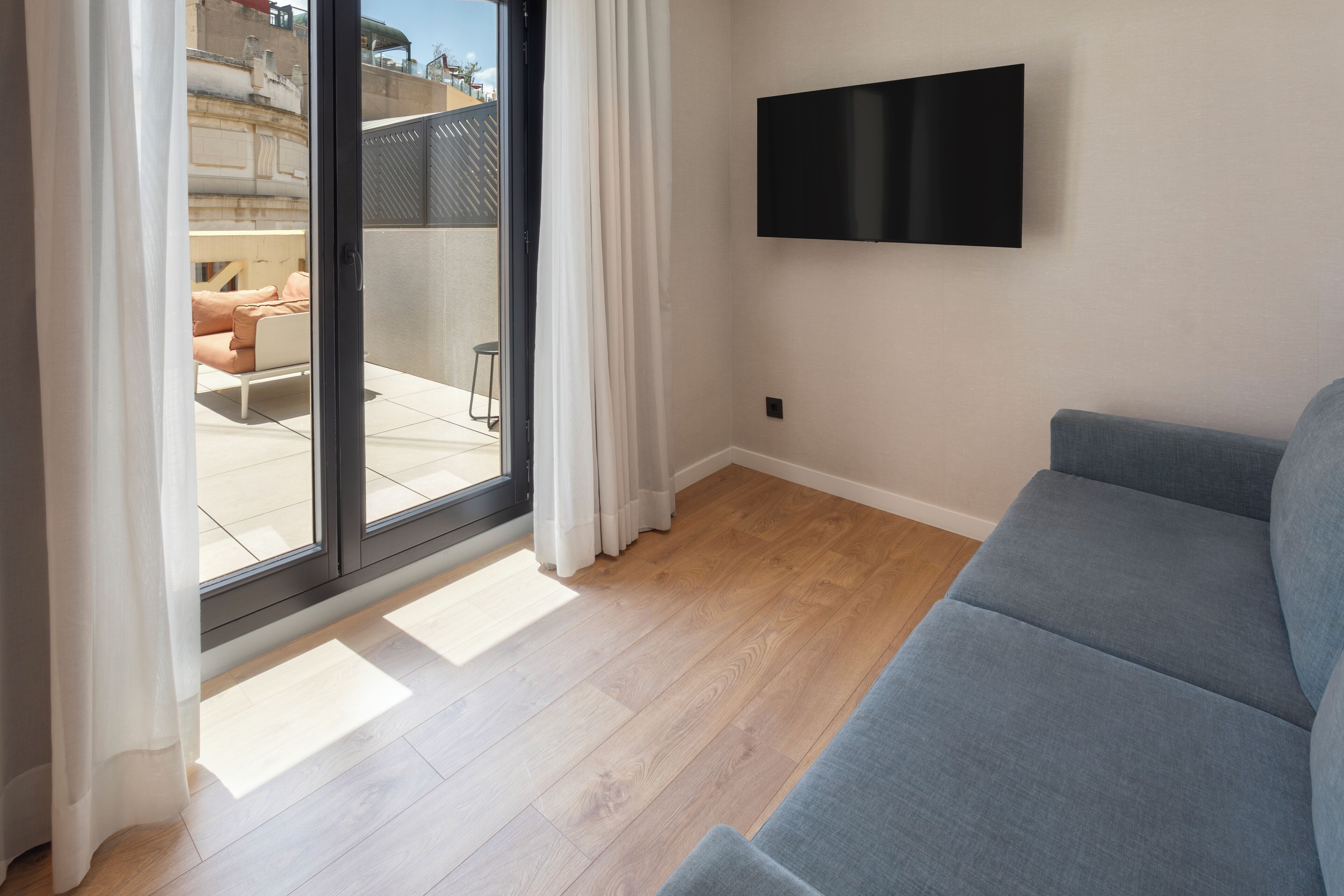A modern living room with a grey sofa, wooden flooring, a flat-screen TV mounted on the wall, and a sliding glass door leading to an outdoor seating area.