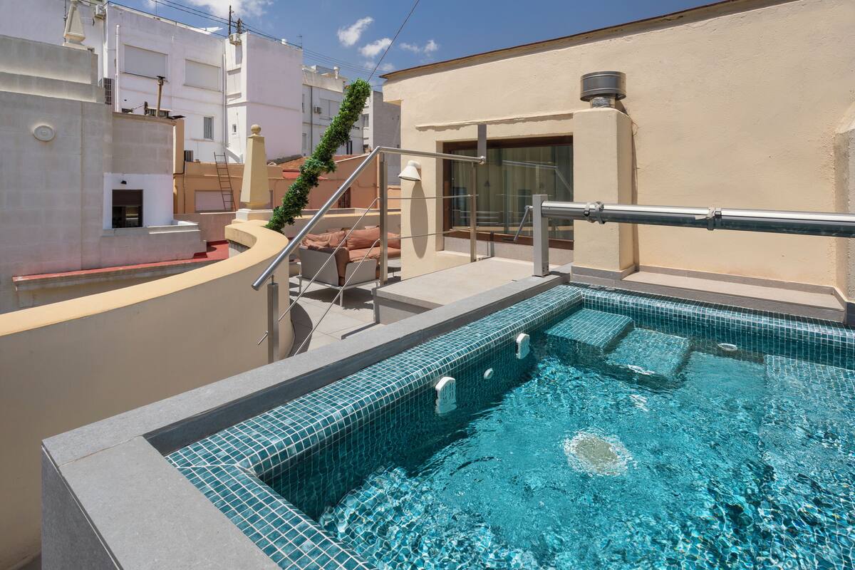 A rooftop swimming pool with clear blue water, a metal railing, and a beige building in the background.