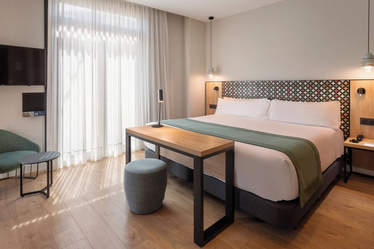 A modern hotel room with a wooden floor, a bed with a patterned headboard, a small table, a chair, and a TV mounted on the wall.