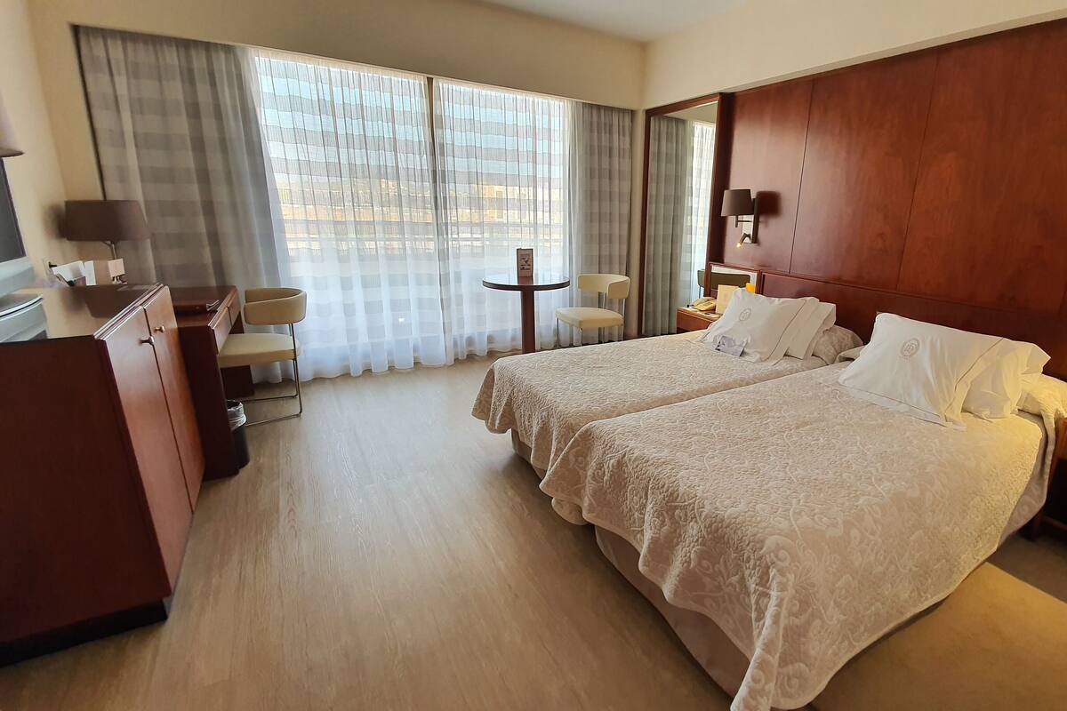 Een hotelkamer met twee bedden, een bureau, een stoel, een kleine tafel en een raam met gordijnen.