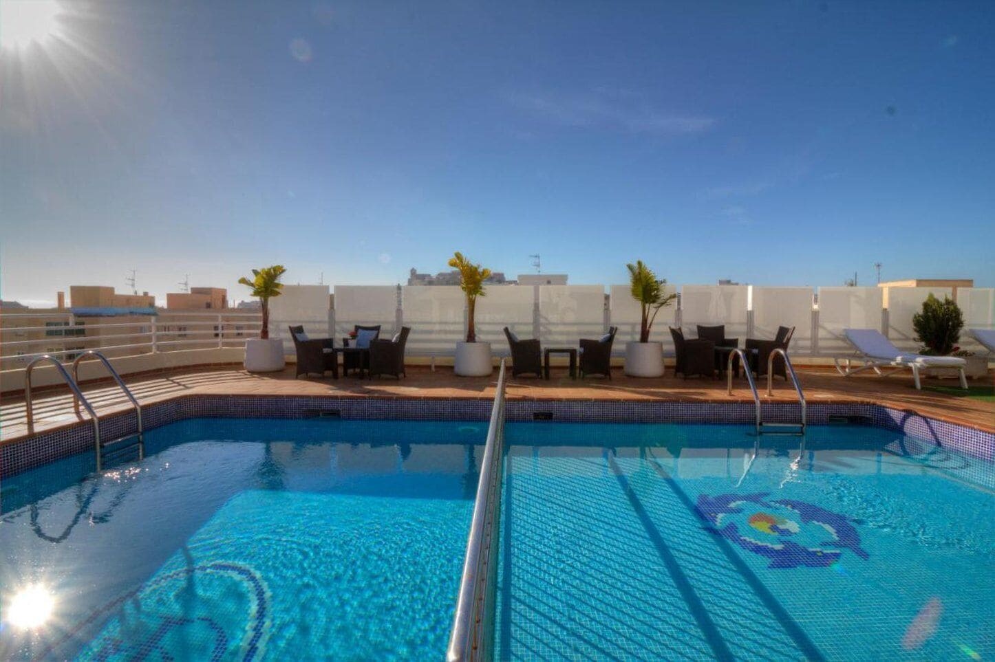 A rooftop swimming pool with lounge chairs and tables, surrounded by a white fence and palm trees.