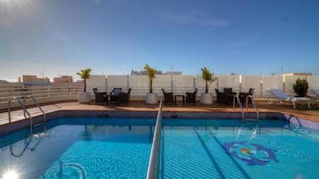 Una piscina en la azotea con sillas de descanso y mesas, rodeada por una valla blanca y palmeras.