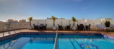 A rooftop swimming pool with lounge chairs and tables, surrounded by a white fence and palm trees.