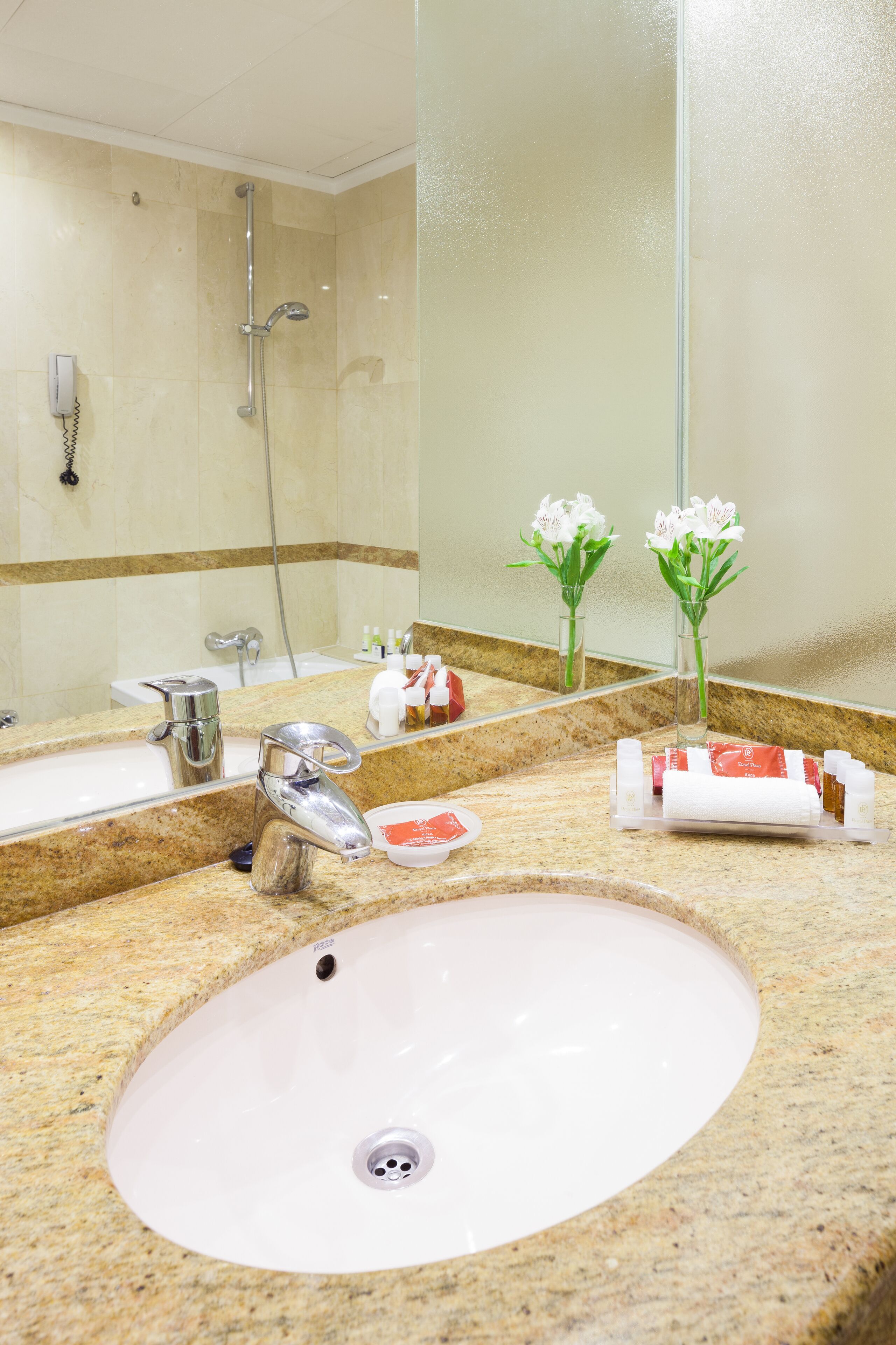 Baño de hotel con lavamanos, espejo, ducha y artículos de aseo.