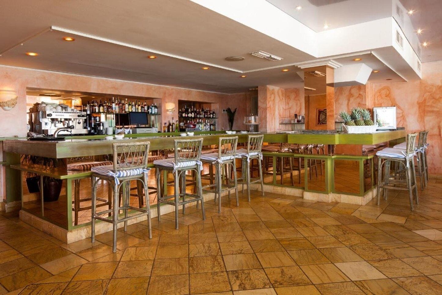 Zona de bar con una barra larga, varios taburetes y una variedad de botellas y vasos en estantes.