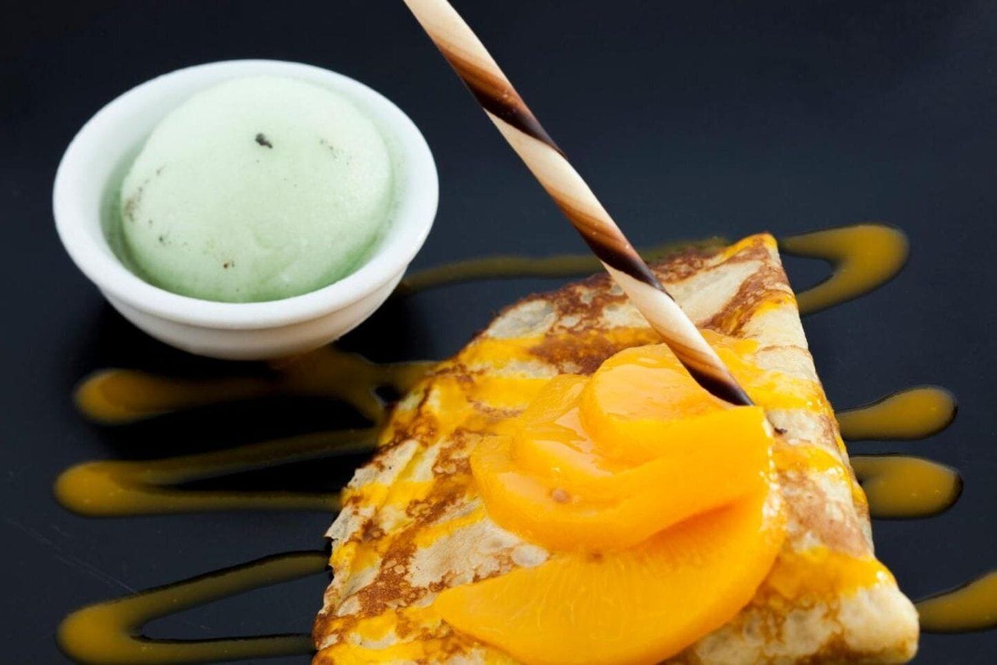 A dessert plate with a crepe topped with caramelized fruit and a small bowl of green ice cream.