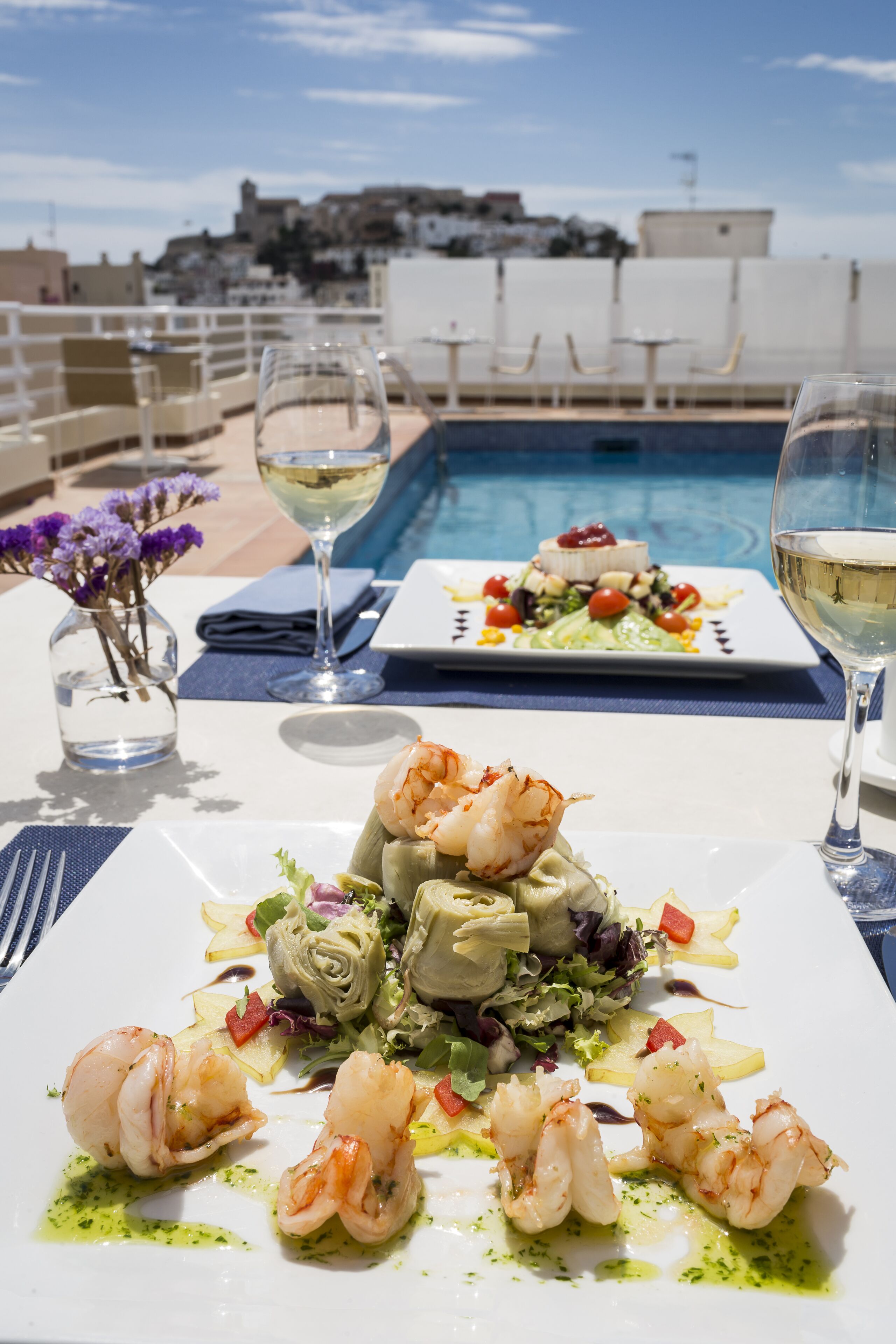 A dining table with seafood dishes, a vase with flowers, and two glasses of white wine.