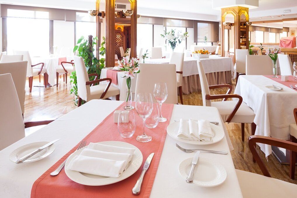 Un comedor con mesas dispuestas para cuatro, cada una con mantel blanco, posavasos rojos y servilletas blancas dobladas con cuidado.