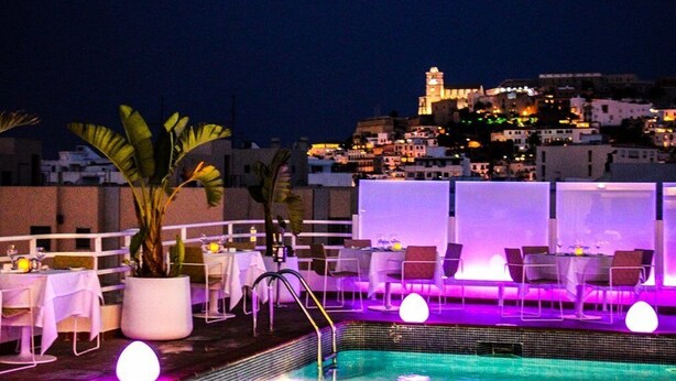 Un área de piscina en la azotea con sillas de descanso y mesas, una piscina y vistas a un paisaje urbano nocturno.