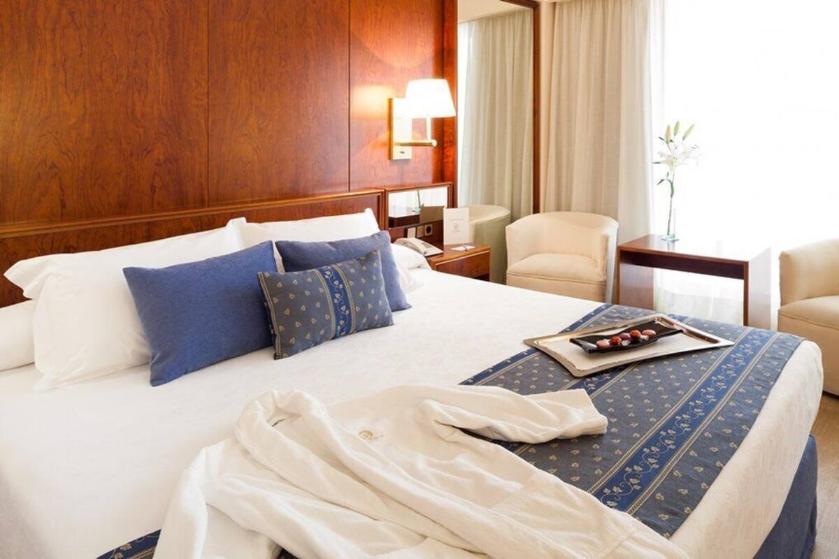 Een hotelkamer met een groot bed, een nachtkastje met een lamp, een stoel en een vaas met bloemen.