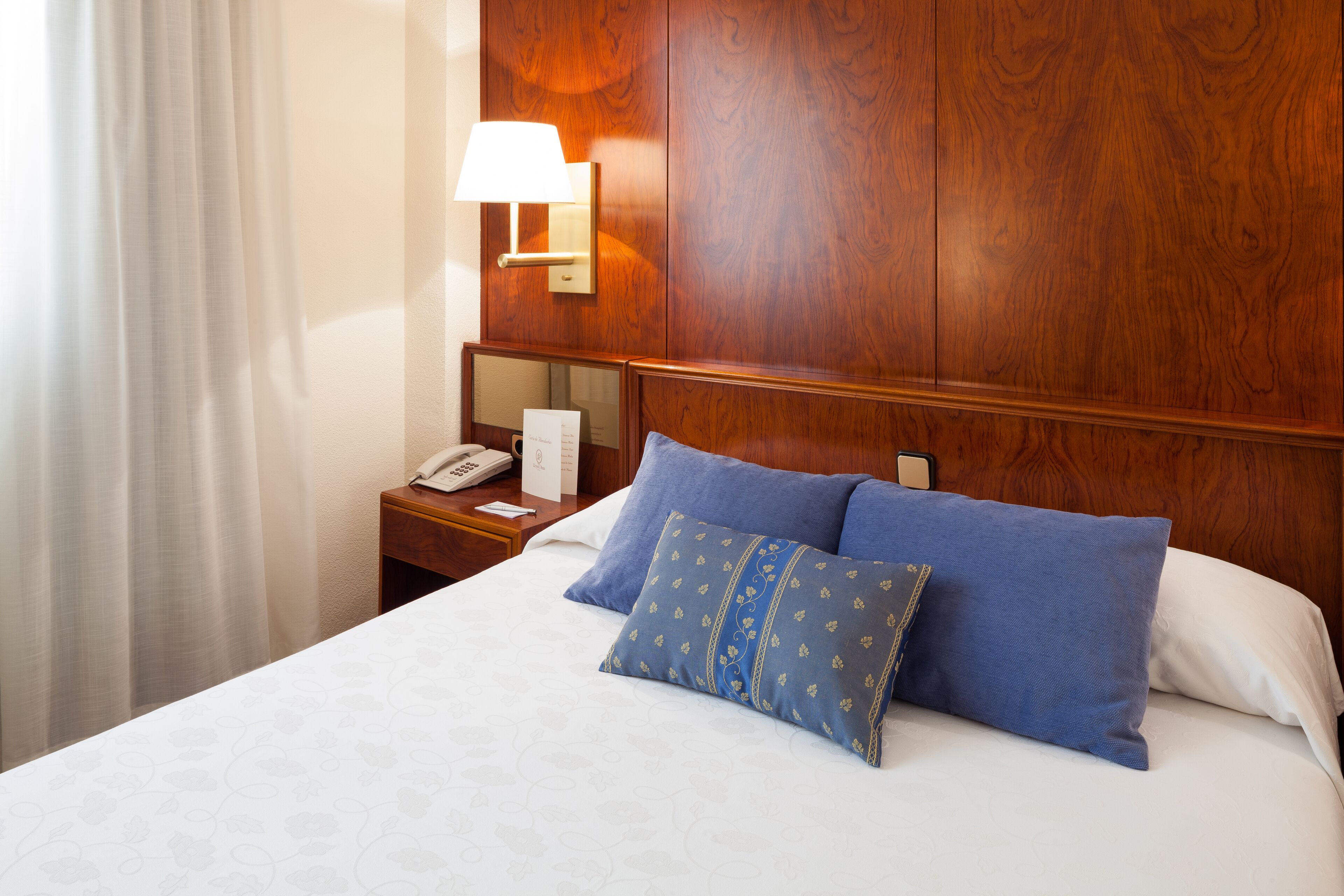A hotel room with a wooden headboard, a bedside table with a lamp, a telephone, and two pillows.