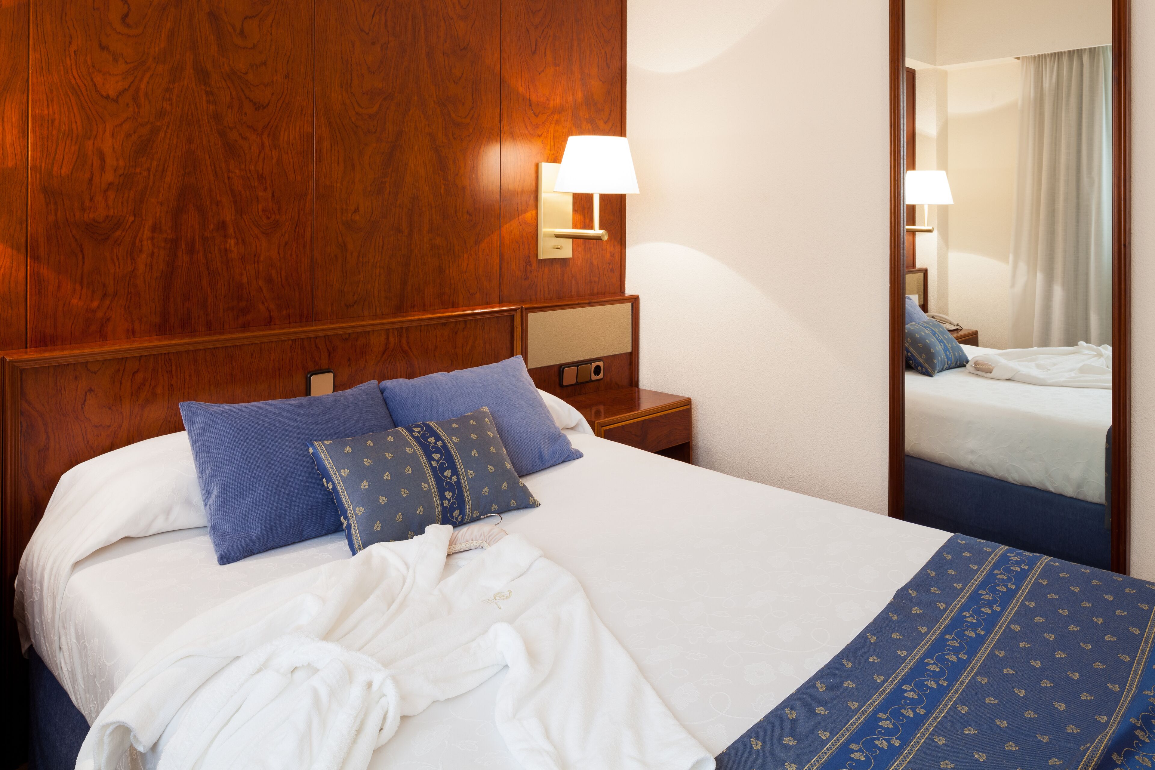A hotel room with a wooden headboard, a large bed with white and blue bedding, a bedside table with a lamp, and a mirror reflecting the room.