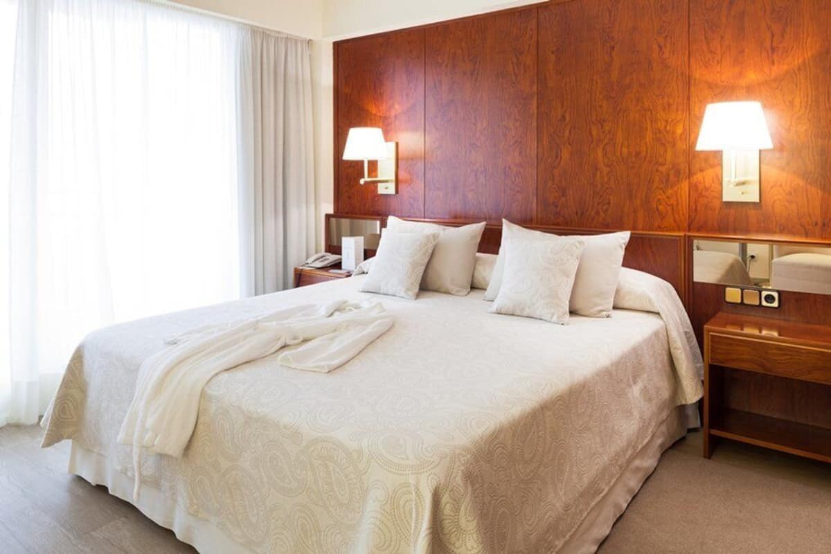 Een hotelkamer met een groot bed, een houten hoofdeinde, twee nachtkastjes en een bijzettafeltje met een lamp.