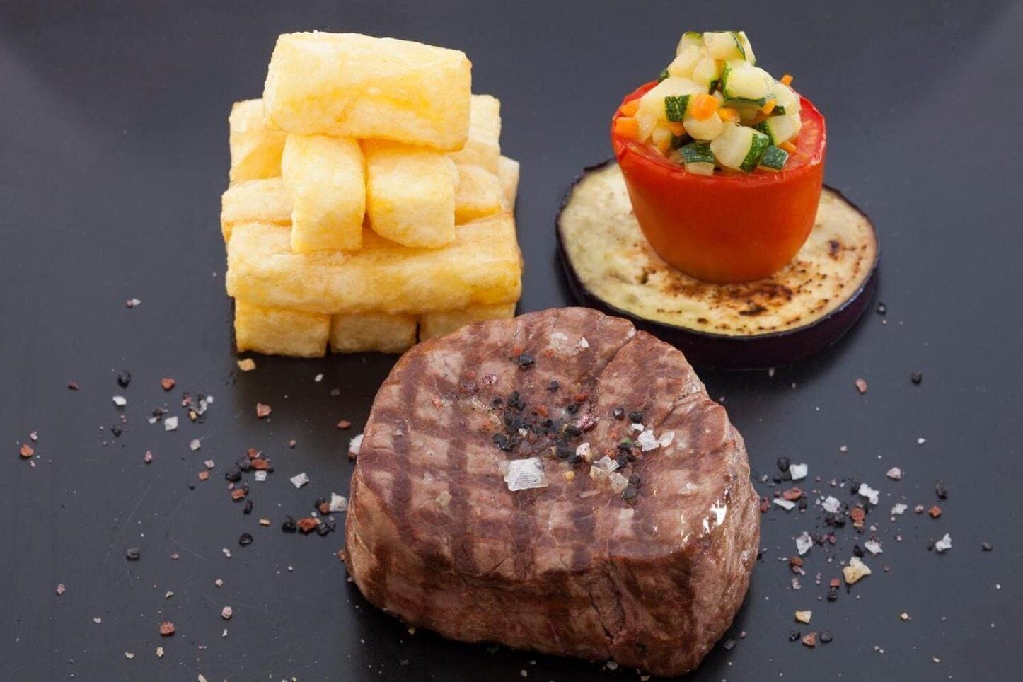A steak, French fries, and a stuffed tomato on a dark plate.