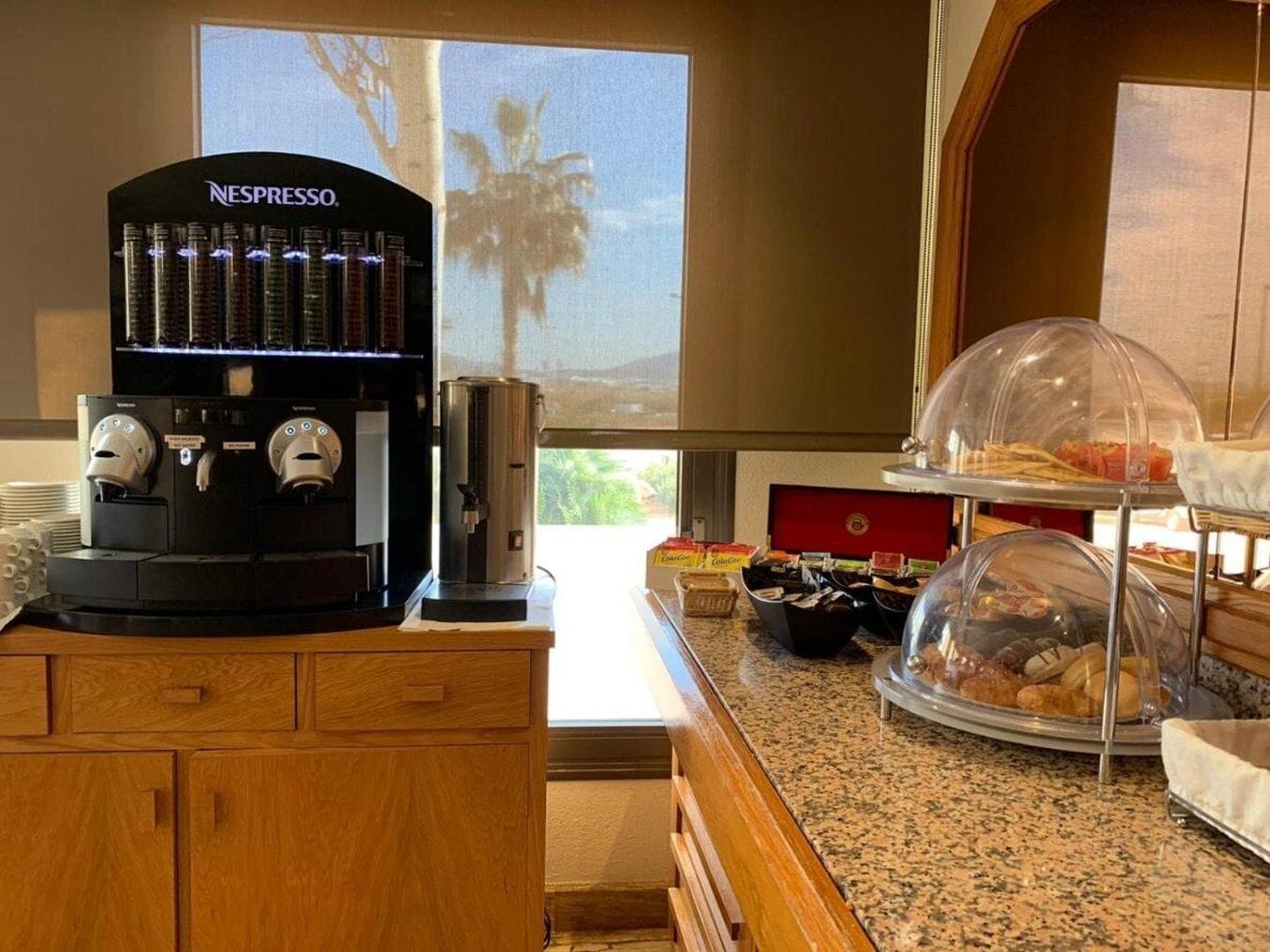 A Nespresso coffee machine, a buffet with pastries, and a view of palm trees through a window.