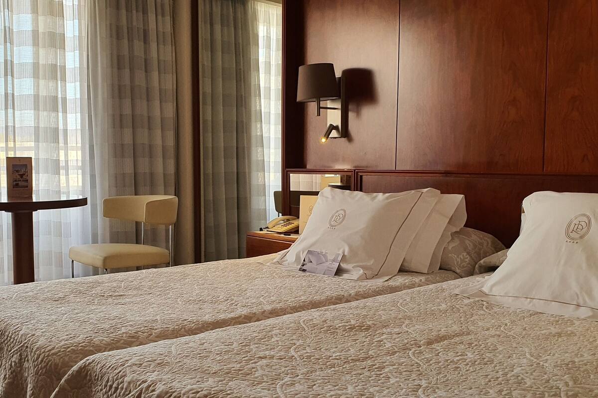 Een hotelkamer met twee bedden, een houten hoofdbord, een nachtkastje, een lamp, een stoel en een balkon met uitzicht op bomen.
