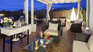 Un patio al aire libre con mobiliario de mimbre, una mesa de vidrio con bebidas y vista a la ciudad al anochecer.