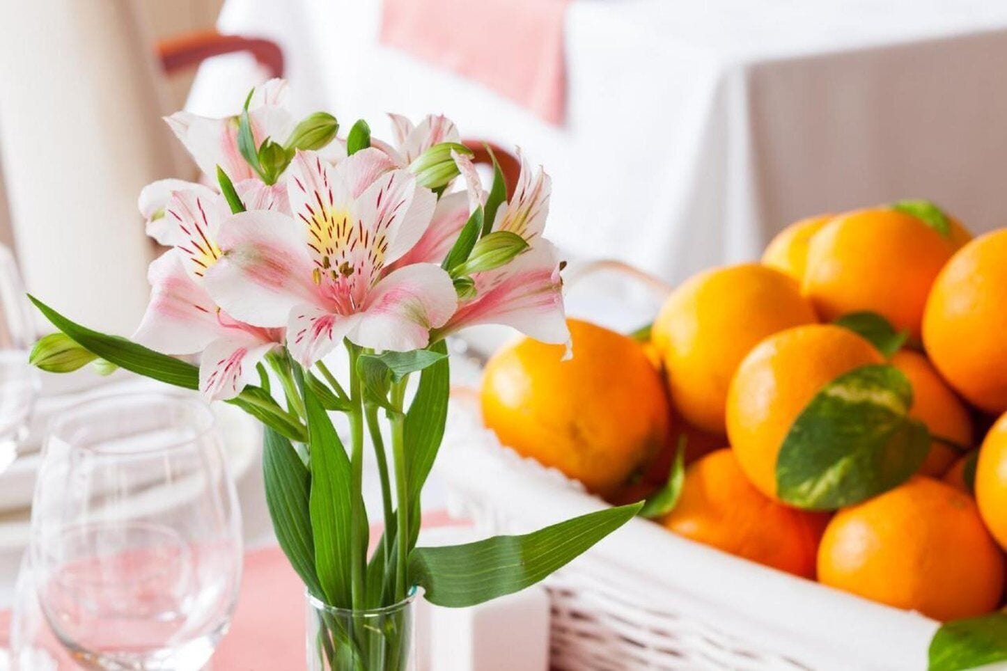 Une table avec un vase de lys roses, un verre d’eau et un panier d’oranges.