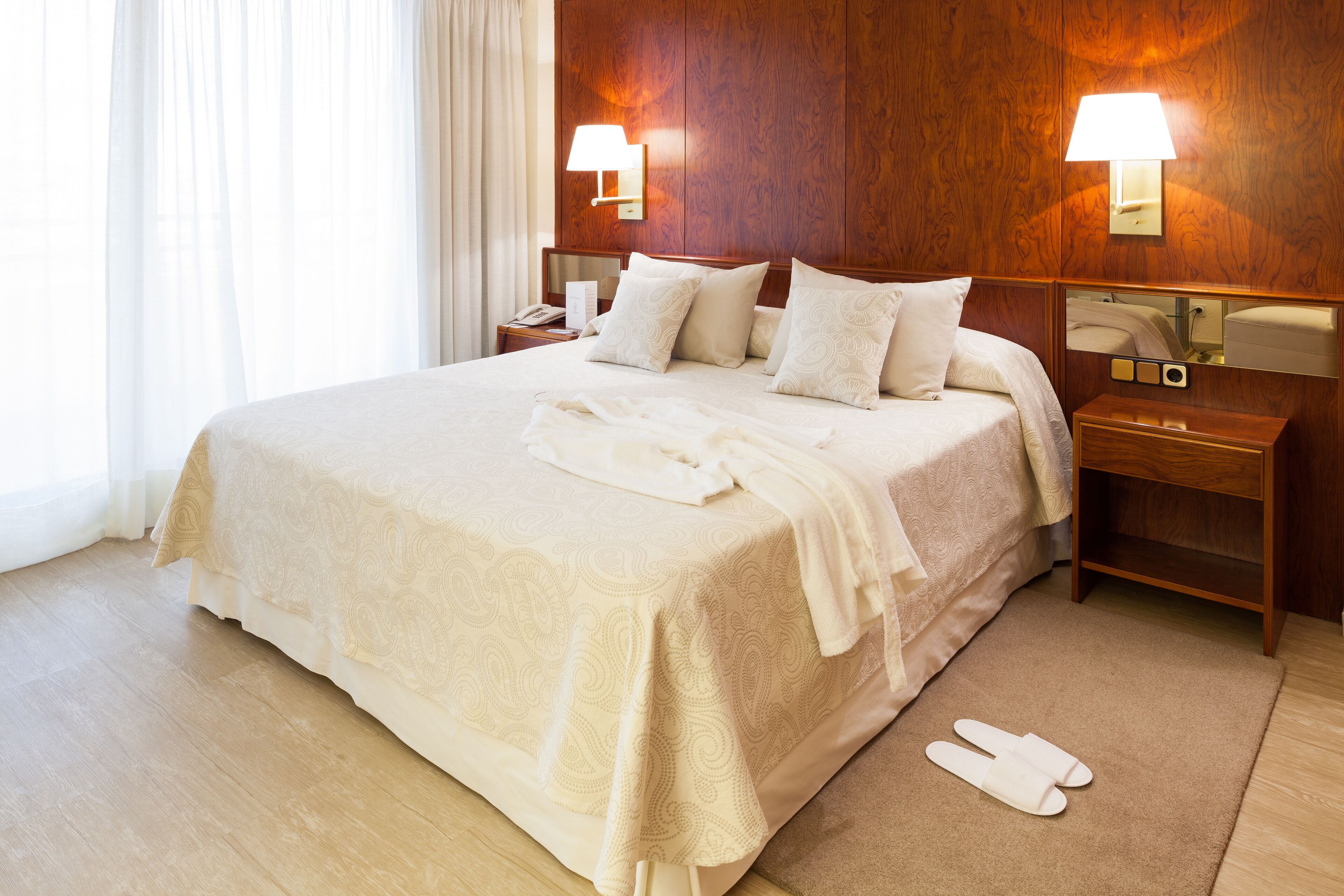 A neatly made bed with a patterned bedspread, two bedside lamps, a nightstand, and a mirror on the wall.