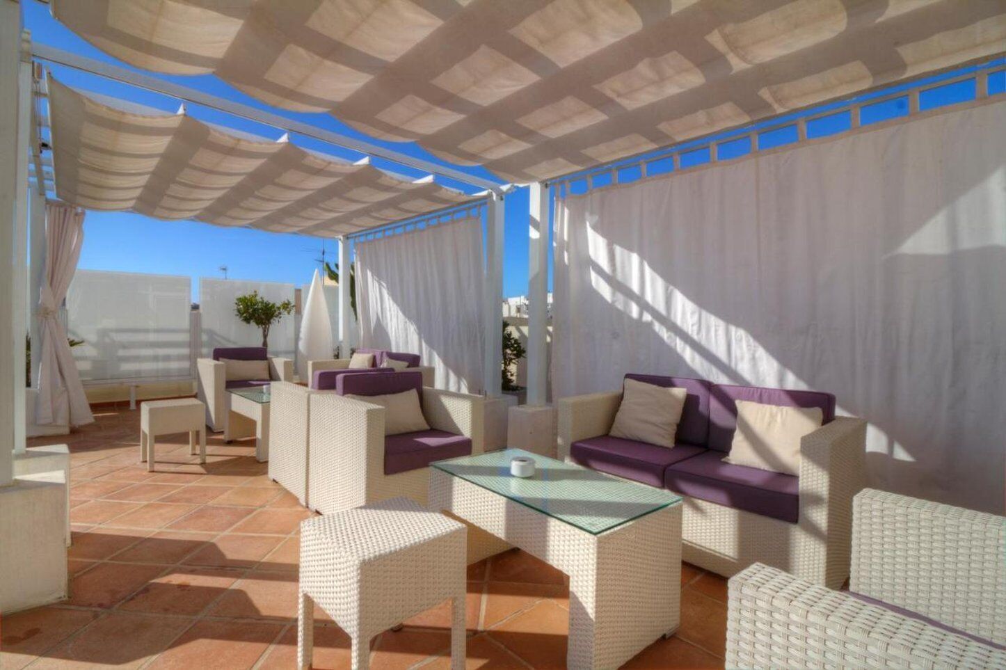 A covered outdoor seating area with wicker furniture and a glass table, featuring a clear view of the sky.