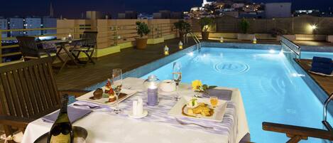A rooftop pool area with a dining setup, including a table with a bottle of champagne, a candle, and a plate of food.