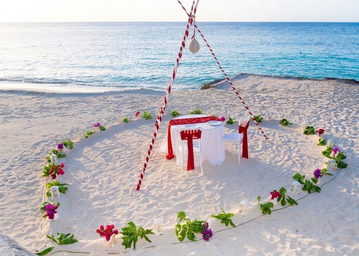 A beach setting with a white table and chairs, surrounded by a red and white striped rope, and decorated with flowers.