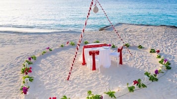 A beach setting with a white table and chairs, surrounded by a red and white striped rope, and decorated with flowers.